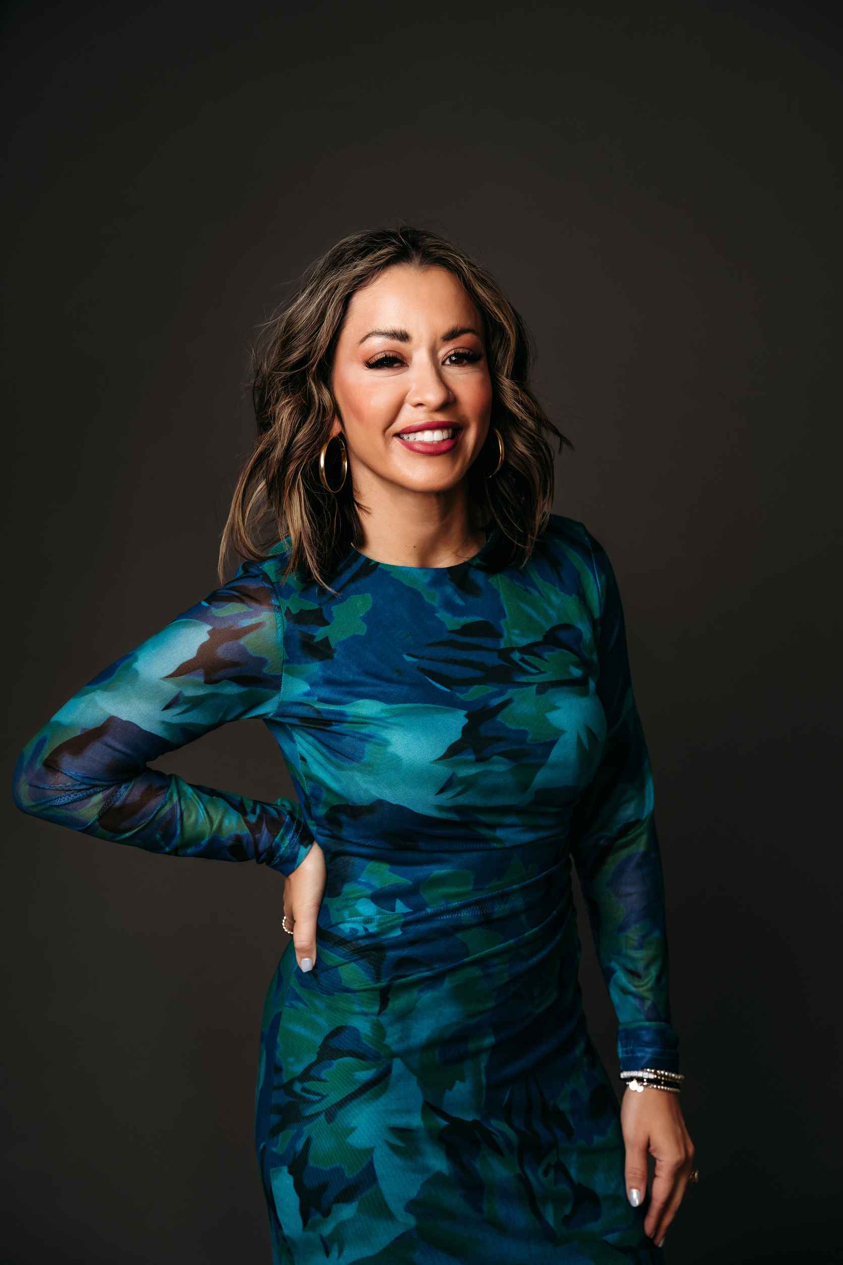 Lexi Yoo NP in a fitted blue and green floral long-sleeve mesh dress, hand on hip, against a dark studio backdrop.