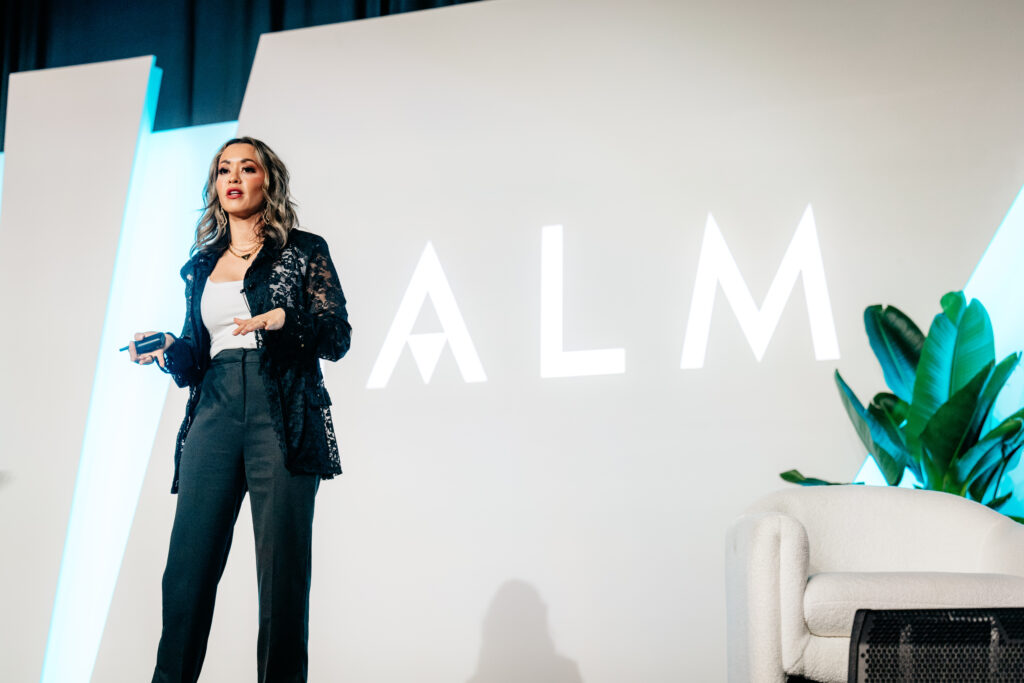 Lexi Yoo NP delivering a keynote at CALM in a navy lace blazer and dark trousers, white CALM-branded stage wall behind her.