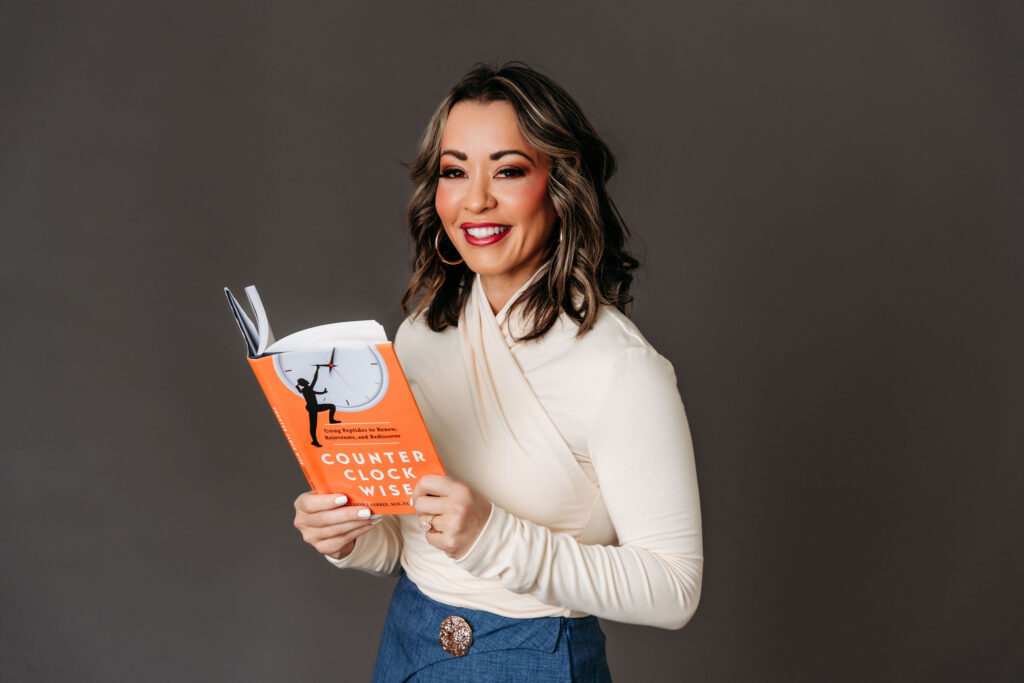 Lexi Yoo NP in a cream wrap top and blue skirt holding the book 'Counter Clockwise: Using Peptides to Renew, Rejuvenate, and Rediscover.'
