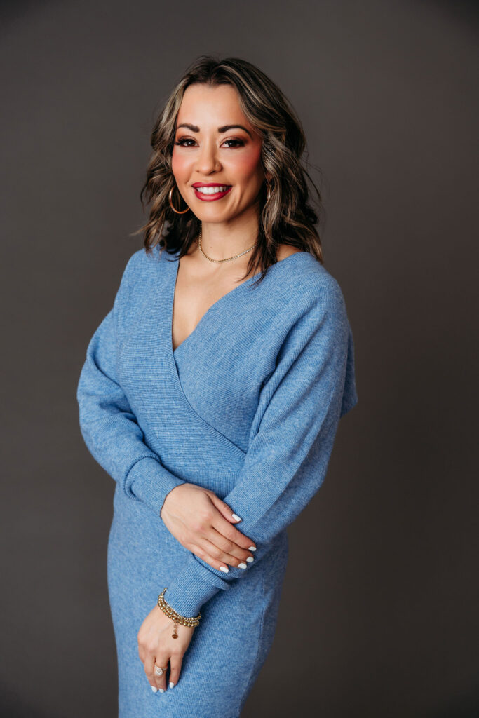 Lexi Yoo NP smiling in a blue knit wrap dress, hands clasped in front, dark studio backdrop.