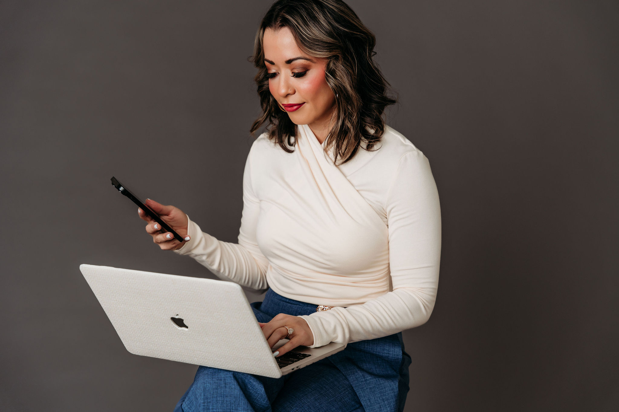 Lexi Yoo NP in a studio portrait reviewing content on her laptop and phone