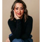 Lexi Yoo, nurse practitioner - Black long-sleeve top, seated on blue stool, hand resting on cheek, tan background - relaxed general portrait