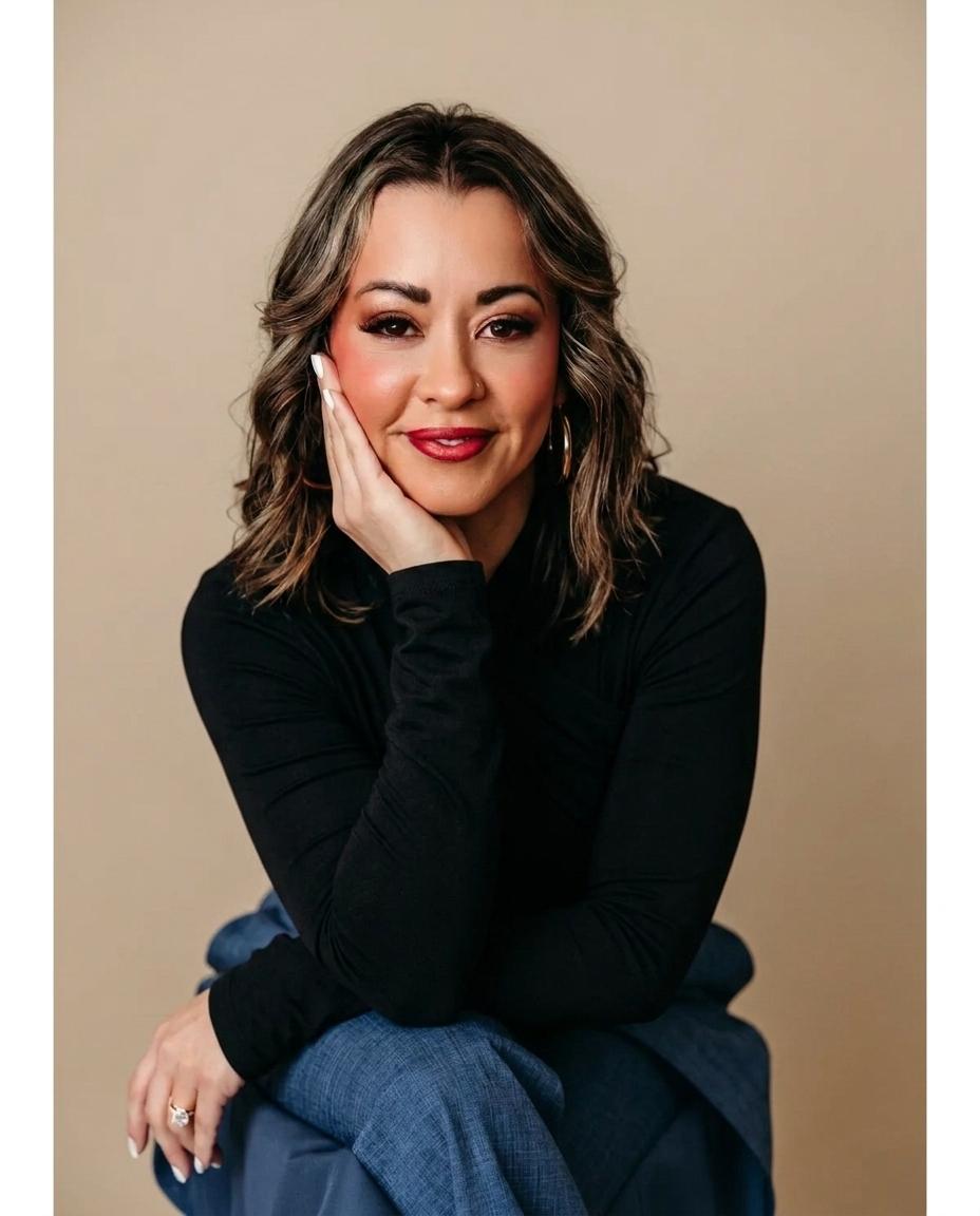 Lexi Yoo, nurse practitioner - Black long-sleeve top, seated on blue stool, hand resting on cheek, tan background - relaxed general portrait