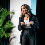 Lexi Yoo, nurse practitioner - On-stage speaking shot - black lace jacket + white top + black pants, mid-gesture, potted plant + stage lighting