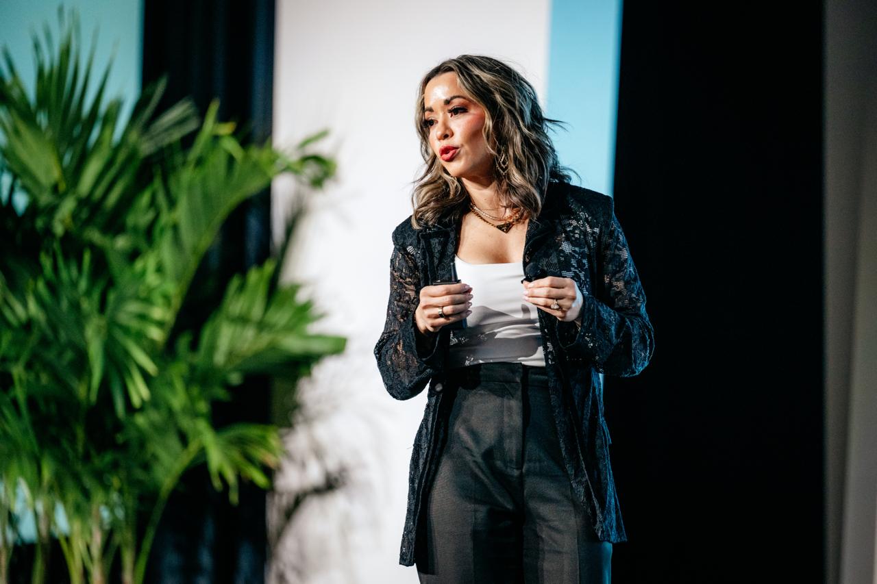 Lexi Yoo, nurse practitioner - On-stage speaking shot - black lace jacket + white top + black pants, mid-gesture, potted plant + stage lighting