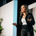 Lexi Yoo, nurse practitioner - On-stage keynote shot - black lace jacket + white top + dark pants, holding a clicker, white stage panels