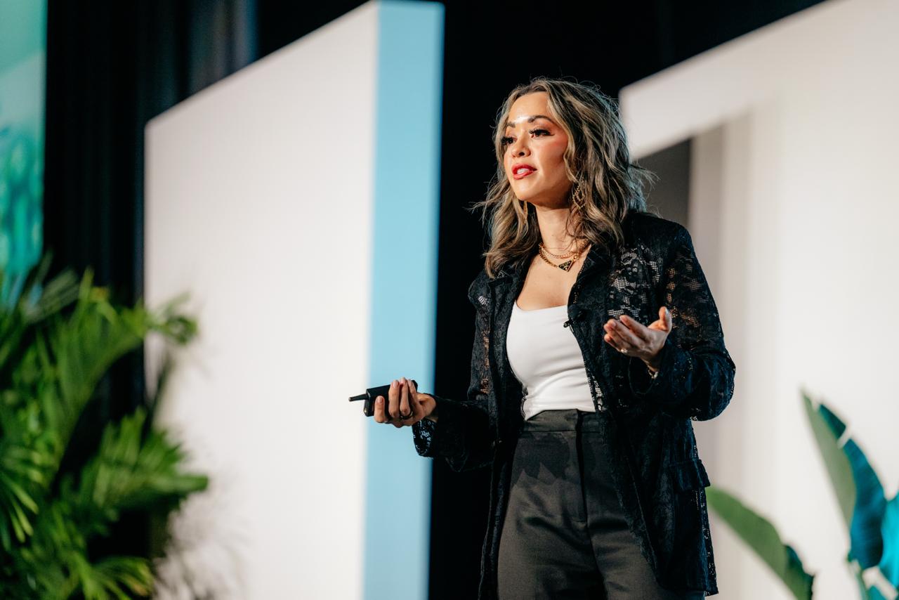 Lexi Yoo, nurse practitioner - On-stage keynote shot - black lace jacket + white top + dark pants, holding a clicker, white stage panels